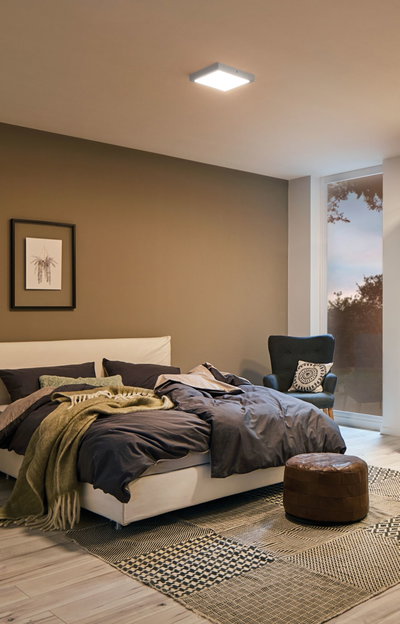 Chambre moderne avec plafonnier carré blanc, lit confortable avec linge de lit foncé et pouf en cuir marron