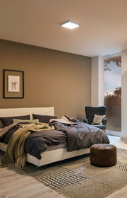 Modernes Schlafzimmer mit weißem Bett, grauer Bettwäsche, braunem Lederhocker und Deckenleuchte