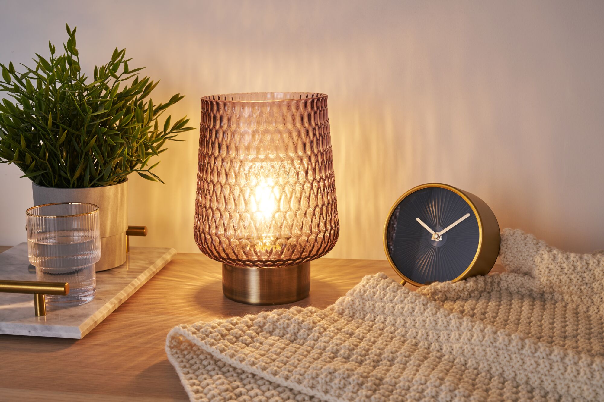 Table lamp with textured pink glass and metal base beside green plant and black clock on wooden surface