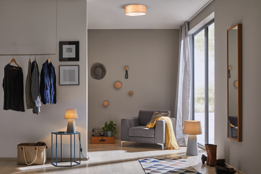 Moderner Wohnraum mit grauem Sessel, Holz-Deckenleuchte und Tischlampe, helle Einrichtung und Fenster.