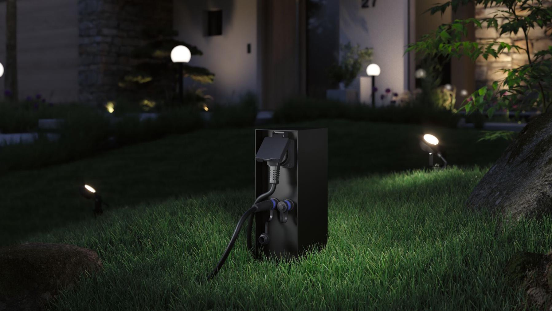 Schwarze Außensteckdose mit Schutzklappe auf Rasen, umgeben von Gartenbeleuchtung und Hausfassade bei Nacht.
