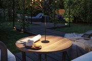 Schwarze LED-Tischleuchte aus Metall auf Holztisch im Garten mit Zeitung und Tasse Kaffee.