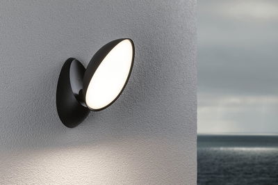 Sort LED-væglampe med rund hvid lampe på grå væg, udendørsbelysning ved havet.