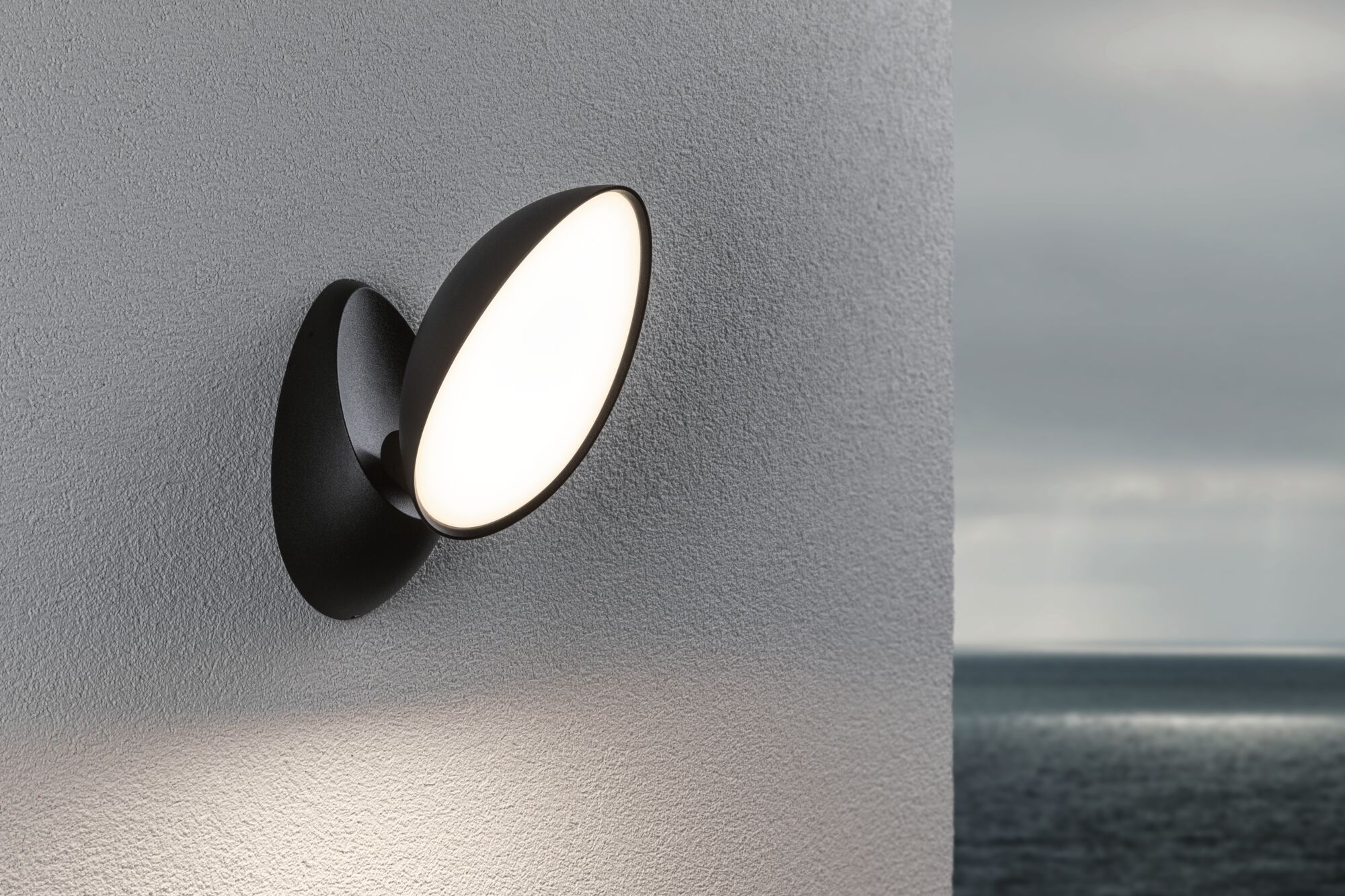 Schwarze LED-Wandleuchte mit rundem, weißem Leuchtmittel an grauer Wand, Außenbeleuchtung am Meer.