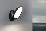 Schwarze LED-Wandleuchte mit rundem, weißem Leuchtmittel an grauer Wand, Außenbeleuchtung am Meer.