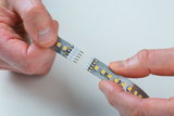 Deux mains connectant une bande LED avec LEDs jaunes et contacts métalliques sur support gris pour éclairage.