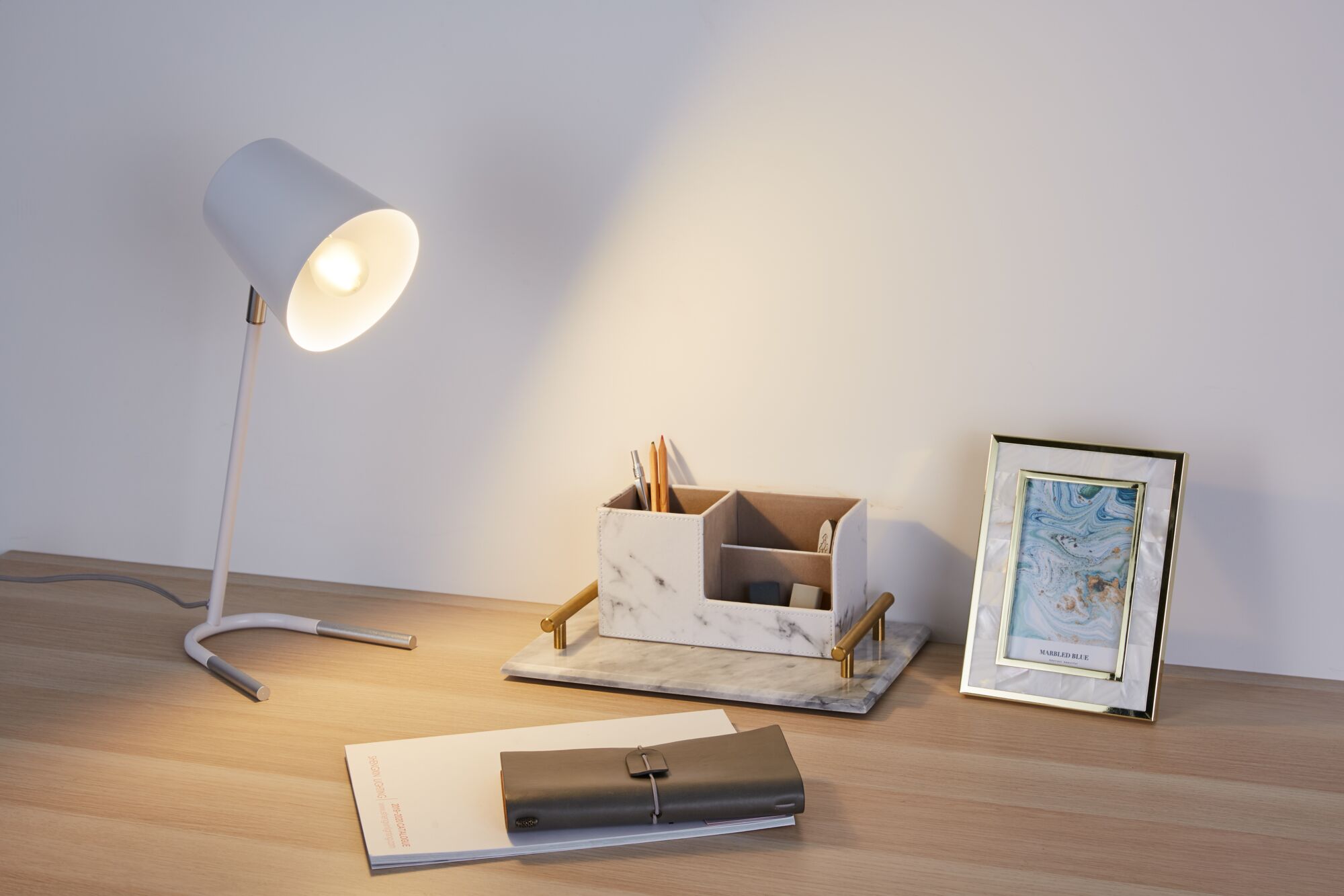 Lampe de bureau blanche avec pied en métal éclairant un bureau en bois avec organiseur en marbre et cadre bleu.