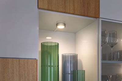 Round white LED under-cabinet light in modern kitchen with green and gray glass vases.