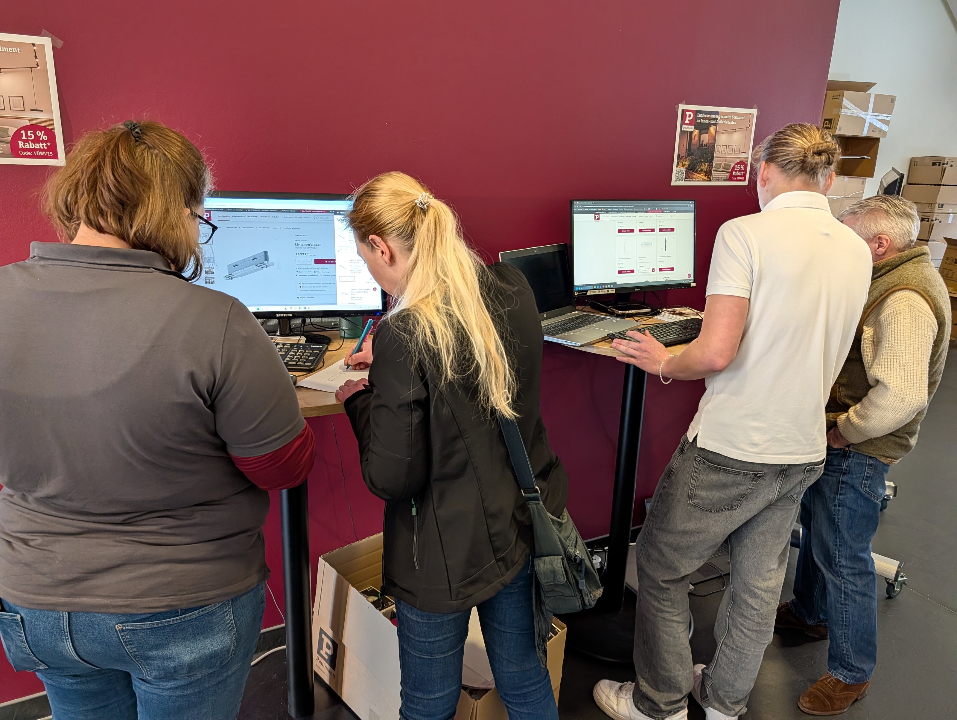 Vier Personen an Stehtischen mit Computern vor roter Wand, Online-Lichtprodukte und Rabattcode sichtbar