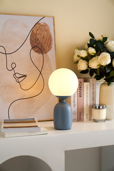 Lampe de table moderne avec base en métal bleu-gris mat et globe en verre blanc sur étagère décorée.