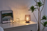 Lampe de table en verre avec abat-jour marron texturé sur buffet blanc près d'un pichet d'eau et plante