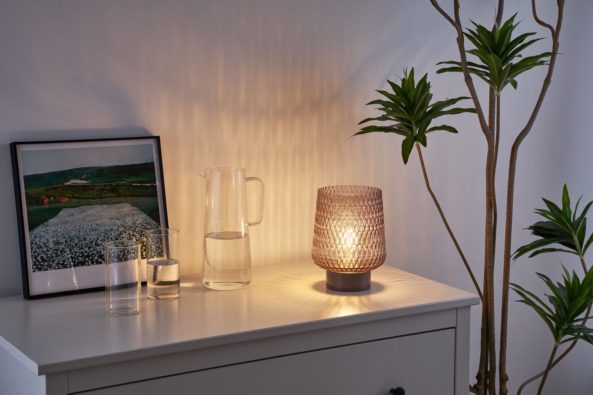 Glass table lamp with textured brown shade on white sideboard next to water jug and plant
