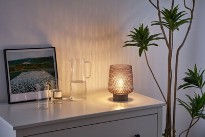 Glass table lamp with textured brown shade on white sideboard next to water jug and plant