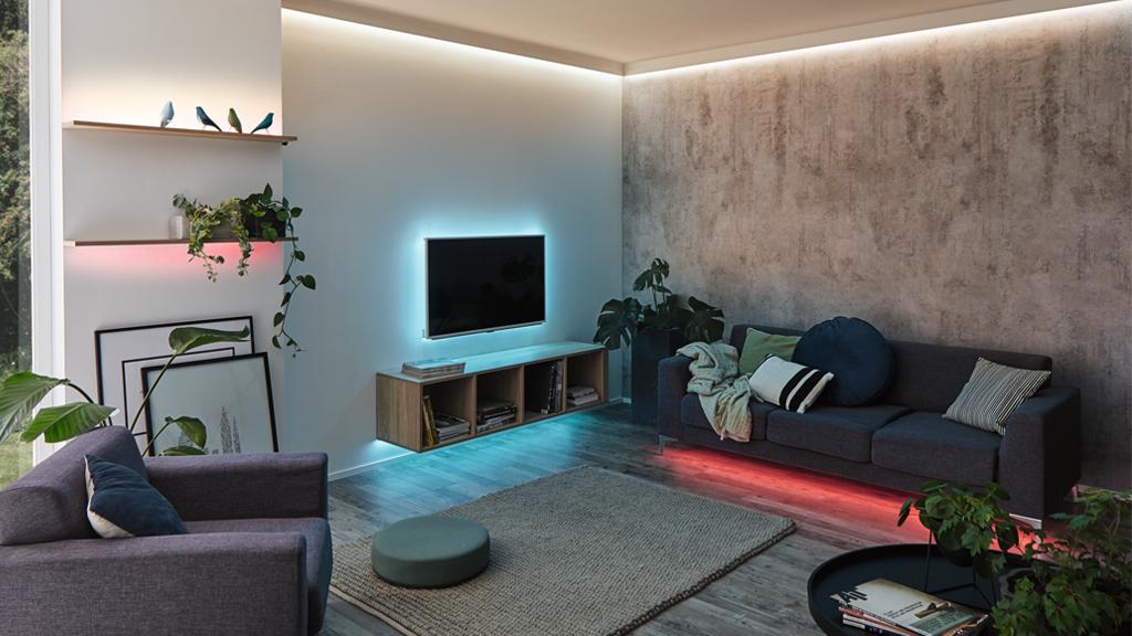 Modernes Wohnzimmer mit LED-Streifen in Blau und Rot, grauem Sofa und Holzregal mit Pflanzen.
