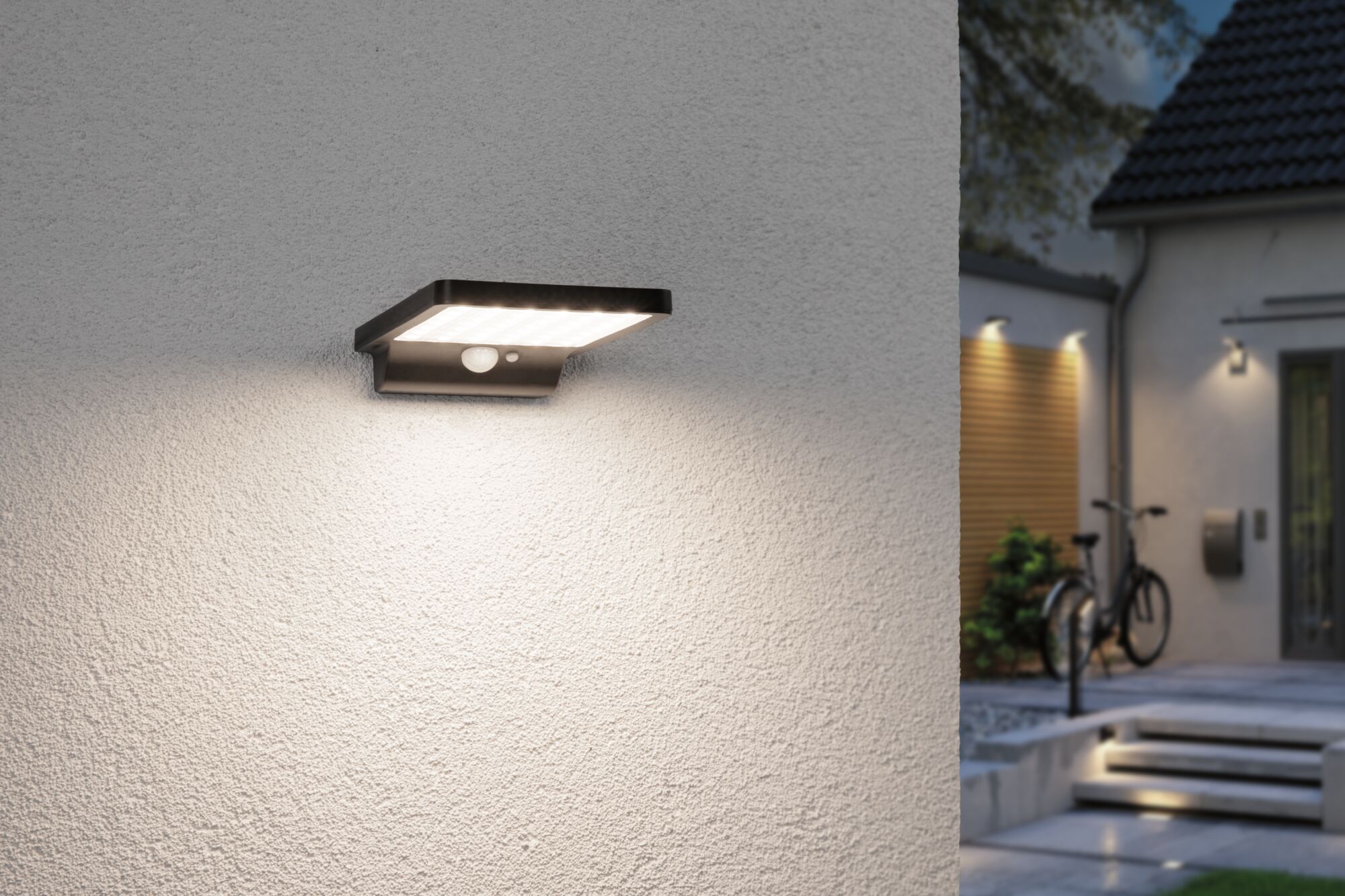 Black LED outdoor wall light with motion sensor mounted on white wall illuminating entrance area.