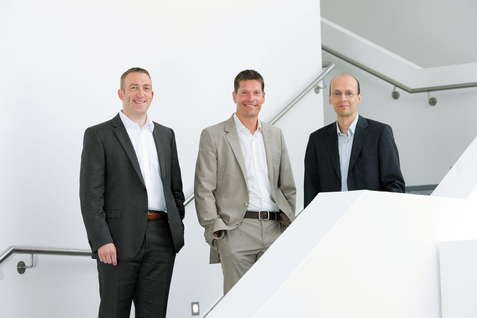 Three men in suits smiling by a white staircase in a modern office setting without product focus.