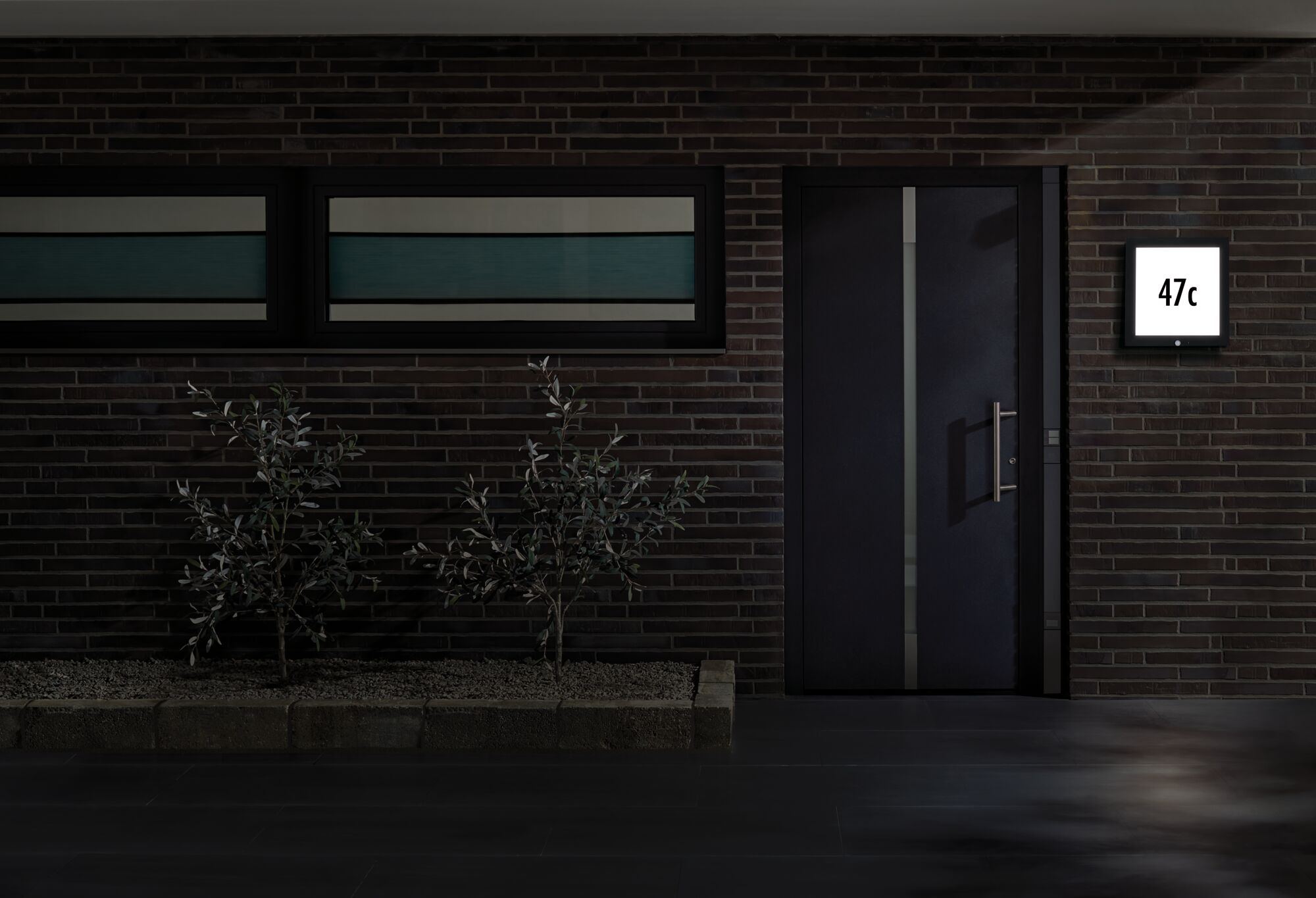 Moderne donkergrijze voordeur met roestvrijstalen handvat en verlicht huisnummerbord aan bakstenen muur.