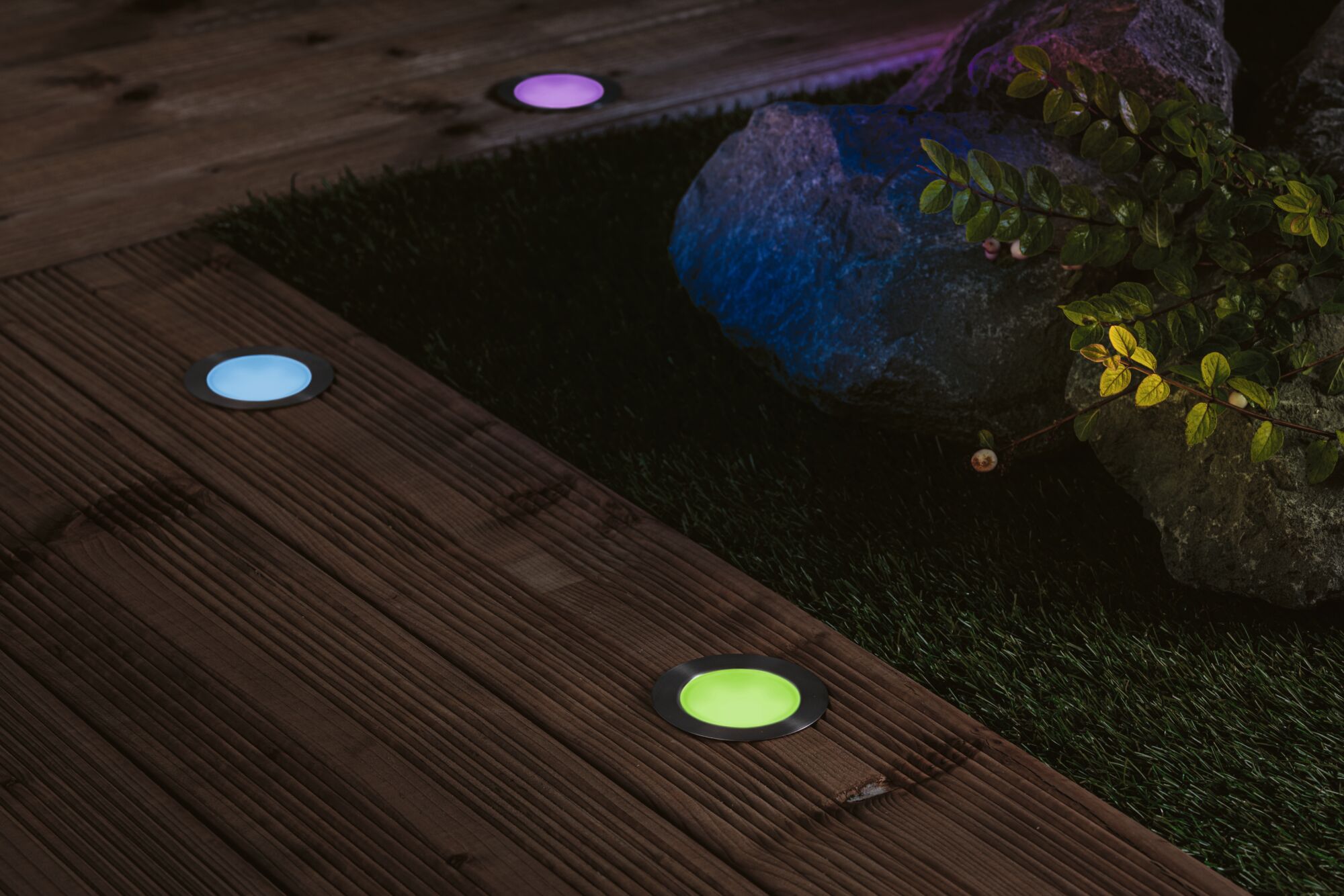 Colorful LED recessed floor lights in stainless steel on wooden terrace by garden with stones and plants