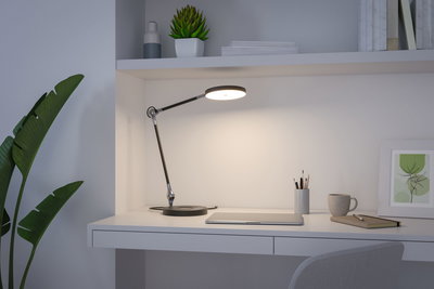 Modern black LED desk lamp with round light head on white workspace with laptop and cup