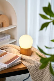 Moderne Tischleuchte mit Holzfuß und milchweißem Glas in gemütlicher Wohnumgebung mit Buch und Decke.