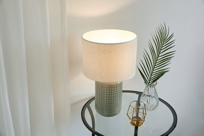 Tafellamp met grijze keramische voet en witte stoffen kap op glazen bijzettafel met decoratie