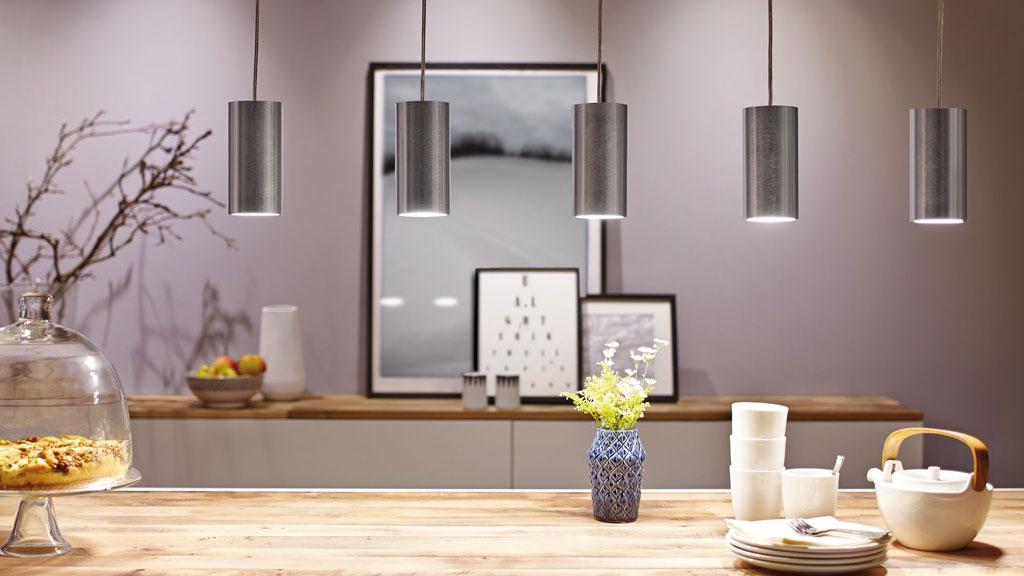 Five silver cylindrical aluminum pendant lights above light wooden table with dishes and vase.