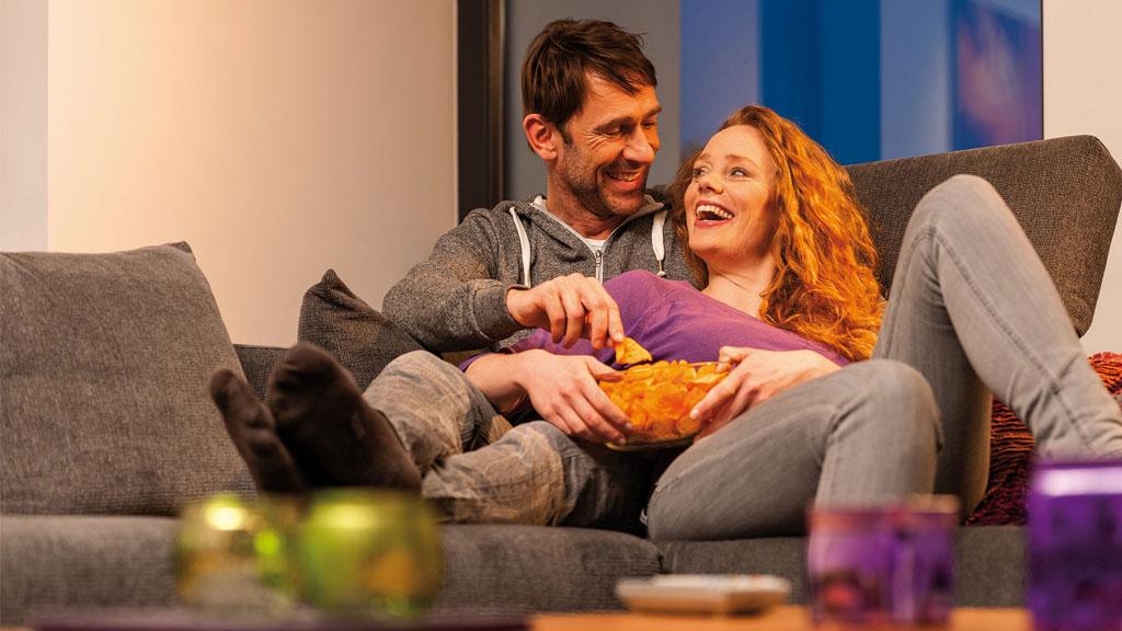 Par slapper af på grå sofa med snacks, hyggelig stue med varmt lys.