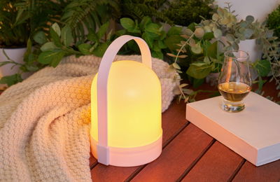 Witte draagbare LED-tafellamp met warm licht op houten tafel naast boek en whiskyglas