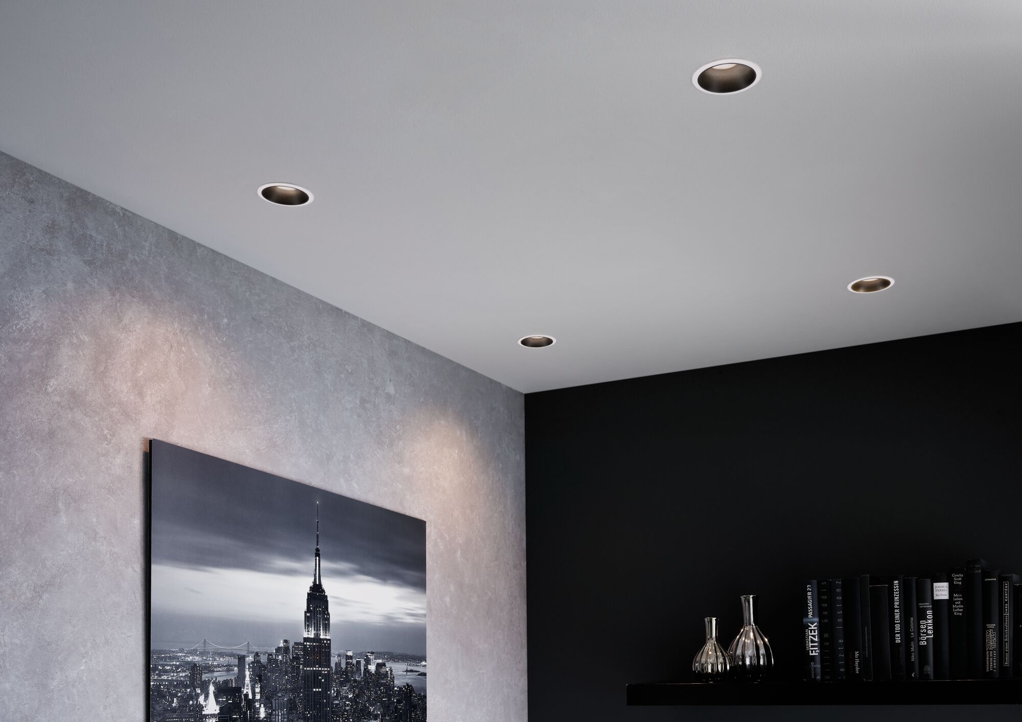 Four round recessed spotlights in white ceiling illuminate modern living room with grey wall and black shelf.