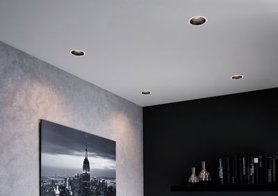Four round recessed spotlights in white ceiling illuminate modern living room with grey wall and black shelf.