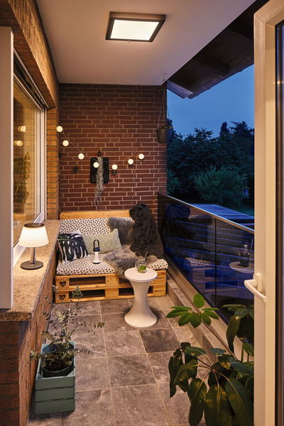Cozy balcony with modern black ceiling light emitting warm white light, decorated with plants and pallet bench.