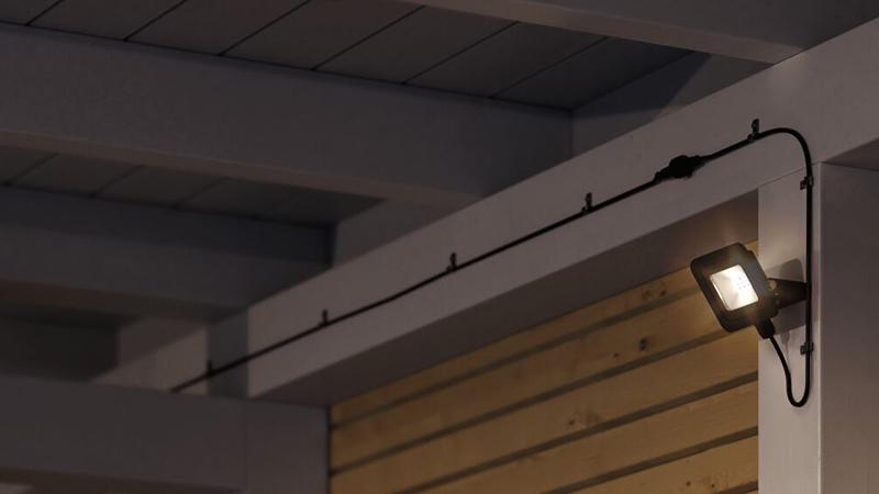 Zwarte LED-buitenwandlamp gemonteerd op witte balk met zichtbare kabel voor tuinverlichting.