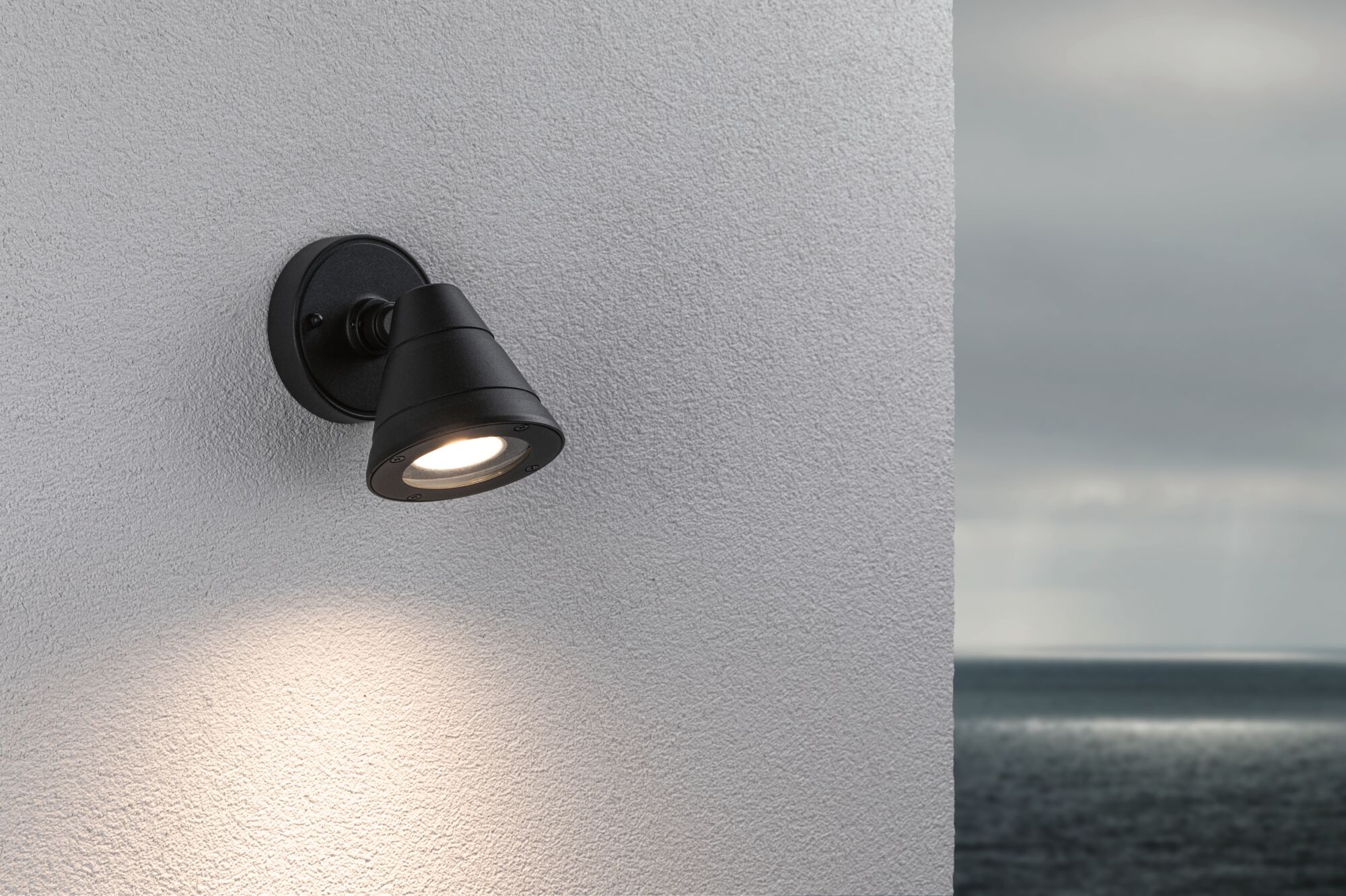 Black metal outdoor wall light with warm white LED on white wall overlooking the sea