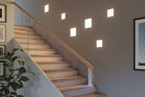 Modern staircase with wooden steps and glass railing, illuminated by square warm white wall lights.