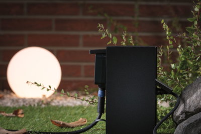 Boîtier extérieur noir avec câble devant une lampe sphérique lumineuse dans un jardin avec herbe et pierres