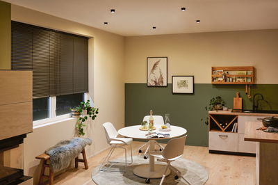 Coin repas moderne avec table ronde blanche, chaises, sol en bois, mur vert-beige et spots au plafond