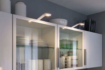Armoires de cuisine blanches à portes vitrées éclairées par des spots LED modernes avec vaisselle et verres.