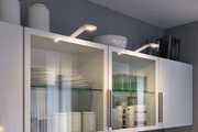 Weiße Küchenschränke mit Glasfront, beleuchtet von modernen LED-Spots, zeigen Geschirr und Gläser.