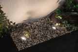 Two round silver LED ground lights on gravel with plants and wall lighting in garden setting