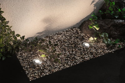 Two round silver LED ground lights on gravel with plants and wall lighting in garden setting