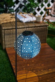 Blue metal solar lantern with decorative pattern hanging on black stand for outdoor garden lighting.