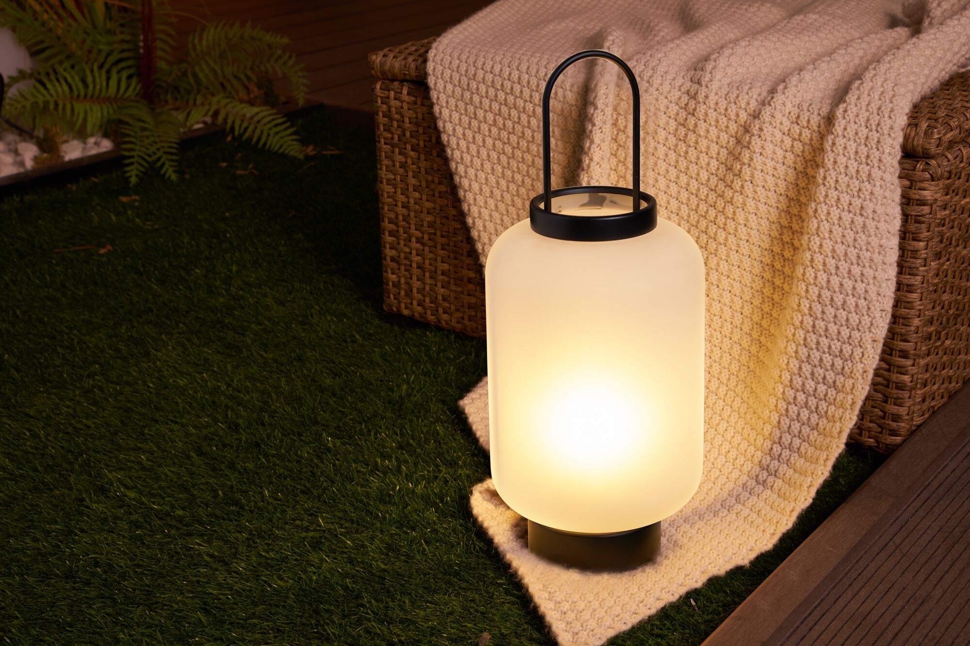Moderne Outdoor-Laterne aus mattweißem Glas mit schwarzem Griff auf grünem Rasen neben Korbsessel.