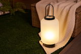 Modern outdoor lantern with matte white glass and black handle on green grass by wicker chair.