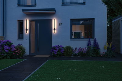 Moderne huisentree met donkere deur en twee slanke wandlampen die warm wit LED-licht uitstralen buiten.