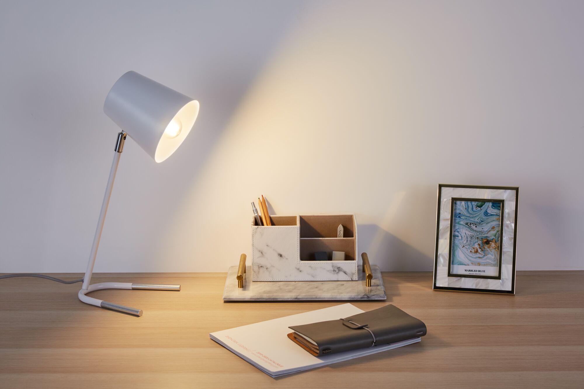 Hvid metal skrivebordslampe med varmt lys på træbord ved siden af marmor organizer og billedramme