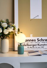 Moderne tafellamp met groene metalen voet en ronde witte glazen lamp naast vaas met witte rozen.