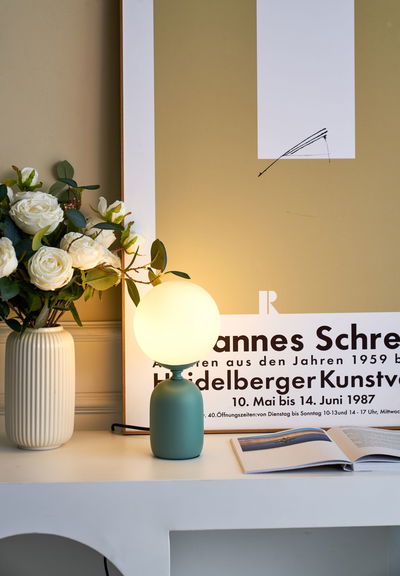 Moderne tafellamp met groene metalen voet en ronde witte glazen lamp naast vaas met witte rozen.