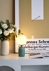 Modern table lamp with green metal base and round white glass light next to vase with white roses.