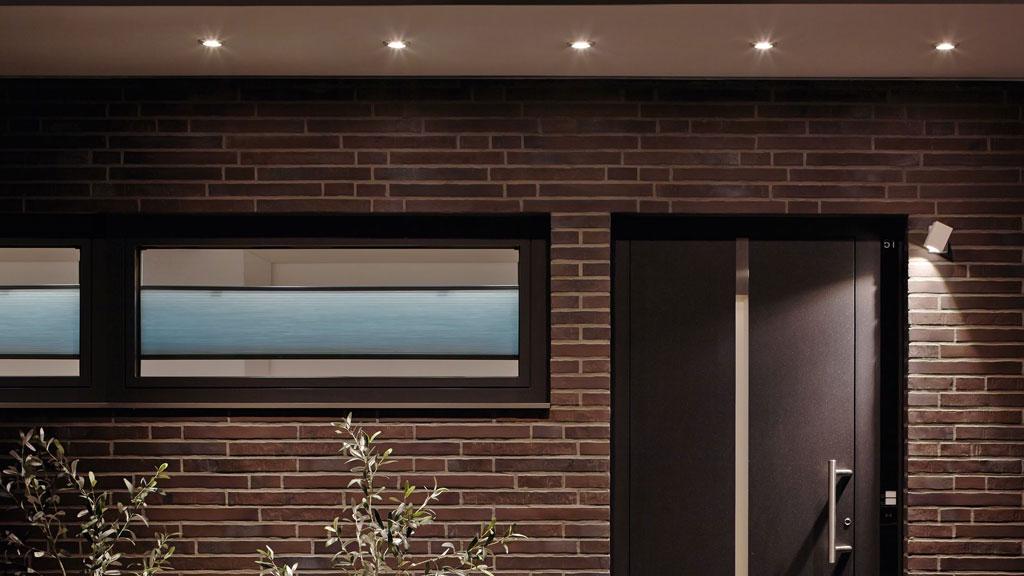 Moderne udendørsbelysning med varm hvide LED-spots på rød murstensfacade og mørk hoveddør.