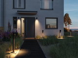 Modernes Haus mit schwarzer Eingangstür und warmweißen Außenleuchten aus Metall am Weg und an der Fassade.